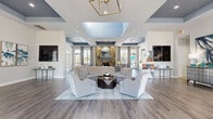 Located at 600 River Birch Ct in Clermont, Florida, this single-story clubhouse offers a generously sized, open-concept layout designed for both comfort and sophistication. The main gathering area is anchored by a dramatic floor-to-ceiling stone fireplace, serving as a striking focal point surrounded by carefully curated seating arrangements that invite conversation and relaxation. Overhead, distinctive coffered ceilings accented in deep navy blue add architectural depth and visual interest, complemented by a geometric brass chandelier that adds a refined, modern touch.

Rich hardwood-style flooring flows continuously throughout the space, tying together the various zones seamlessly. Abundant natural light pours in through large, elegantly draped windows, offering views of the surrounding Florida landscape. Multiple seating vignettes furnished with stylish upholstered chairs and sofas create versatile areas for gathering.

A dedicated office or leasing area features glass-paned French doors, providing a professional yet welcoming environment. The space also includes a well-appointed kitchenette outfitted with gray cabinetry, dark granite countertops, light blue subway tile backsplash, and modern appliances. Contemporary artwork in blue and neutral tones is displayed throughout, reinforcing the cohesive, polished aesthetic of this impressive community facility.