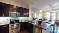 Located in Dallas, TX, this single-story residence at 4601 Amesbury Dr offers a thoughtfully designed interior that blends modern aesthetics with functional living. The kitchen is a standout feature, showcasing rich dark espresso cabinetry with glass-front uppers, stainless steel appliances including a range, microwave, and dishwasher, a green glass tile backsplash, and sleek dark countertops. Pendant lighting illuminates the kitchen island and dining area, which flows seamlessly into the open-concept living space via warm hardwood flooring. The living room is bright and welcoming, with large floor-to-ceiling windows and glass doors that invite generous natural light and offer views of the surrounding neighborhood. The primary bedroom features plush carpeting, a bold accent wall, and ample window light, while the secondary bedroom is equally comfortable and well-lit. The bathroom is elegantly finished with a wide vanity, framed mirror, tile flooring, a glass-enclosed walk-in shower, and direct access to a spacious walk-in closet with built-in shelving and rod storage. Throughout the residence, the design maintains a cohesive contemporary style with clean lines and quality finishes.