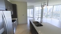 This single-floor residence showcases a thoughtfully designed open-concept layout that seamlessly connects the kitchen and living areas. The kitchen serves as the centerpiece, featuring a large island with a light quartz countertop, a double undermount sink with a contemporary arc faucet, and dark cabinetry that contrasts elegantly with the bright surroundings. Pendant lighting hangs above the island, adding a refined touch. Stainless steel appliances, including a built-in oven and microwave, are integrated along the kitchen wall. Light-toned hardwood flooring runs throughout, contributing to a warm and cohesive aesthetic. The living area is bathed in abundant natural light, courtesy of expansive floor-to-ceiling windows that frame dramatic, unobstructed views of the surrounding urban skyline. A cylindrical column near the window wall adds an architectural element to the space. The overall ambiance is clean, contemporary, and airy, with neutral tones and modern finishes creating a polished environment well-suited to urban living.