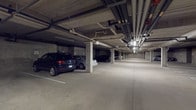 This facility encompasses two distinct functional areas: a covered parking garage and a dedicated fitness center. The parking garage features a structured concrete design with clearly marked stalls, load-bearing support columns, and an extensive network of overhead utility pipes and conduits running along the ceiling. Overhead lighting illuminates the space efficiently, and the smooth concrete flooring accommodates multiple vehicles with ease.

The fitness center is thoughtfully designed to support a range of exercise routines. The room features durable dark flooring, neutral warm-toned walls, and flush ceiling lighting that ensures a well-lit workout environment. A full-length mirrored wall panel expands the sense of space and allows users to monitor their form during exercise. The room is equipped with cardio machines including treadmills and an elliptical trainer, as well as a multi-functional cable strength machine. Exercise balls are available for stretching and core training. A wall-mounted television provides entertainment during workouts, while windows fitted with blinds allow for natural light. Practical amenities include a stainless steel drinking fountain, a paper towel and sanitizer dispenser, a thermostat, and a waste bin, ensuring the space remains clean and comfortable for all users.