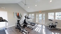 Located at 6100 Barrowood Dr in Raleigh, NC, this single-floor fitness center is thoughtfully designed to provide a bright, motivating environment for exercise. The space is anchored by soaring vaulted ceilings that create an open, airy atmosphere, complemented by a modern ceiling fan that ensures comfortable air circulation throughout workouts. Recessed lighting and an abundance of natural light streaming through multiple windows and a set of elegant French doors illuminate the rich, hardwood-style flooring below. The cardio equipment area is well-appointed, featuring elliptical trainers, a treadmill, and additional machines arranged to allow for comfortable movement between stations. The French doors open directly to an outdoor area, offering both ventilation and a seamless connection to the exterior. Colorful wall art adds a welcoming touch to the otherwise clean, neutral aesthetic defined by soft gray walls and crisp white trim. The overall ambiance is clean, modern, and functional, making this fitness center a practical and inviting amenity for residents of the Barrowood Drive community in Raleigh.