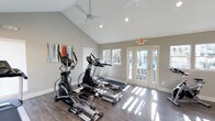 Located at 6100 Barrowood Dr in Raleigh, NC, this single-floor fitness center is thoughtfully designed to provide a bright, motivating environment for exercise. The space is anchored by soaring vaulted ceilings that create an open, airy atmosphere, complemented by a modern ceiling fan that ensures comfortable air circulation throughout workouts. Recessed lighting and an abundance of natural light streaming through multiple windows and a set of elegant French doors illuminate the rich, hardwood-style flooring below. The cardio equipment area is well-appointed, featuring elliptical trainers, a treadmill, and additional machines arranged to allow for comfortable movement between stations. The French doors open directly to an outdoor area, offering both ventilation and a seamless connection to the exterior. Colorful wall art adds a welcoming touch to the otherwise clean, neutral aesthetic defined by soft gray walls and crisp white trim. The overall ambiance is clean, modern, and functional, making this fitness center a practical and inviting amenity for residents of the Barrowood Drive community in Raleigh.