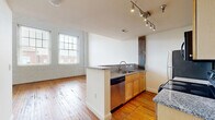 Located on West Broad Street in Richmond, Virginia, this two-story, 3-bedroom, 2-bathroom home showcases a compelling blend of historic industrial character and contemporary finishes. The main floor offers a generous open-plan living and kitchen area anchored by warm, original hardwood floors and striking white-painted exposed brick walls that flood the space with natural light through large multi-pane windows. The kitchen features granite countertops, a central peninsula with a stainless steel sink and dishwasher, light wood cabinetry, and stainless steel appliances, including a range and refrigerator. Rustic exposed wooden beams and columns punctuate both floors, serving as dramatic architectural focal points that nod to the building's history. A staircase with a classic wooden banister leads to the upper level, which houses the bedrooms, including a well-proportioned primary bedroom with two closets offering ample storage. Bedrooms on the upper floor feature hardwood flooring, clerestory windows for natural light, and exposed beam details. Both bathrooms are finished with tile flooring, tub-shower combinations, and wood-toned cabinetry. The two closets provide practical storage throughout the home.
