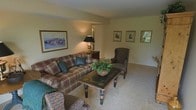 This single-floor living space offers a warm, traditionally styled interior centered around a comfortable seating arrangement. The living room features a plaid-upholstered sofa and coordinating armchair, providing a classic, inviting atmosphere. A decorative coffee table with ornate legs serves as the focal point of the seating area, topped with a lush potted plant that adds a natural touch. Soft, neutral-toned carpeting runs throughout, contributing to the room's cozy ambiance. Warm lighting is provided by a traditional table lamp positioned on a side table near the sofa, casting a gentle glow across the space. The walls are adorned with a collection of framed artwork, including a large landscape print and a series of smaller framed pieces arranged in a gallery-style grouping. The open layout connects seamlessly into an adjacent area, suggesting a spacious and functional floor plan. The overall aesthetic is classic and homey, with traditional furnishings and warm tones creating a welcoming, lived-in feel throughout the space.