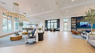Located at 2045 Paseo Belluno in Chula Vista, CA, this single-story community space offers a generously proportioned, open-concept interior designed with contemporary coastal elegance. The entire floor is unified by striking herringbone-patterned light wood flooring, crisp white walls, and soaring tray ceilings fitted with recessed lighting that keeps the atmosphere bright and welcoming throughout.

The main living area is anchored by a bold navy blue built-in shelving unit housing a flat-screen TV, flanked by large windows that frame outdoor views of the surrounding neighborhood and palm trees. A statement gold-toned chandelier adds a refined focal point to this lounge zone, complemented by warm leather accent chairs and a circular coffee table arrangement.

Adjacent to the lounge is a fully equipped kitchen island finished in white marble, with bar seating, sleek black pendant lighting, light wood cabinetry, and a wall-mounted TV—ideal for social gatherings. A cozy navy-framed window nook provides an intimate seating retreat nearby.

A separate entry area with expansive grid-pane windows floods the reception zone with natural light, while curated blue-toned artwork adorns the walls. A dining area with contemporary sculptural seating completes the space. Throughout, the design balances modern sophistication with an inviting, community-focused ambiance.