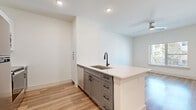 This thoughtfully designed one-bedroom, one-bathroom unit offers a clean, modern aesthetic across its single floor. The open-concept kitchen and living area serves as the heart of the space, anchored by a central kitchen island with a white quartz countertop, an undermount sink, and a matte black faucet. Gray shaker-style cabinetry with black hardware complements the light wood-look flooring that flows throughout. Stainless steel appliances, including a full-size refrigerator and range, complete the functional kitchen. The living area benefits from natural light streaming through a large window, enhancing the bright, airy atmosphere. A dedicated storage nook with built-in open shelving provides convenient organization space. The bedroom is a comfortable retreat, finished with plush carpeting, a ceiling fan, and a sizable window with outdoor views of greenery. A five-panel interior door adds a refined architectural touch. The bathroom is well-appointed with a glass-enclosed walk-in shower featuring tile surround, a gray vanity with quartz countertop, a large framed mirror, and cohesive matte black fixtures throughout. The overall design is cohesive, clean, and contemporary.