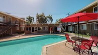 This residential community centers around a generously sized outdoor swimming pool that serves as the focal point of the complex. The pool area is enclosed by a secure metal fence and features a smooth concrete deck lined with lounge chairs, shaded umbrella seating, and a dedicated spa. The clear, turquoise water reflects the open sky, creating an inviting atmosphere on sunny days. A convenience vending machine is conveniently located poolside, adding a practical amenity for residents. The surrounding buildings exhibit a classic mid-century architectural style, clad in warm brown and tan tones with wood-accented facades. Each building offers exterior walkways, individual unit entrances, and private balconies accessible via exterior staircases. The multi-building layout creates a semi-enclosed courtyard feel, with mature trees providing natural shade and softening the built environment. The complex is designed with security and safety in mind, with gated pool access and clearly posted pool guidelines. The overall ambiance is relaxed and communal, well-suited for outdoor leisure and everyday residential living in a sunny California-style setting.