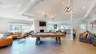 This thoughtfully designed single-floor community clubhouse offers a dynamic and inviting atmosphere ideal for socializing and relaxation. The expansive main gathering area serves as the heart of the space, anchored by a rustic wood-framed pool table positioned beneath a series of striking multi-arm brass chandeliers that add a modern, sophisticated touch. Soaring vaulted ceilings with exposed architectural beams amplify the sense of openness, while light wood-tone flooring runs throughout, tying the space together seamlessly. A textured white brick fireplace with a wall-mounted flat-screen TV creates a warm focal point, flanked by large windows that flood the room with natural light and offer views of the surrounding neighborhood. Multiple seating zones are thoughtfully arranged throughout, including plush leather sectionals, upholstered lounge chairs, and casual conversation groupings accented with colorful throw pillows and curated artwork. A shuffleboard table and high-top bar seating provide additional entertainment options. A cozy sunroom nook features a tufted caramel leather sectional bathed in natural light. A dedicated meeting area includes a round wood dining table with upholstered chairs and a large-scale landscape painting. The kitchenette is equipped with stainless steel appliances, a coffee station, and a decorative patterned tile backsplash, making it ideal for hosting gatherings.