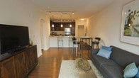 This single-floor space showcases a thoughtfully designed open-concept layout that seamlessly connects the main living areas. Rich hardwood flooring flows throughout, tying together the living room, dining area, and kitchen into one cohesive and inviting environment.

The kitchen is a standout feature, boasting dark espresso cabinetry, granite countertops, and stainless steel appliances, including a full-size refrigerator. A peninsula with bar-height seating and elegant pendant lighting above makes it both functional and stylish. Track lighting runs across the ceiling, providing ample illumination throughout the space.

The dining area, positioned adjacent to the kitchen, accommodates a modern glass-top table with contemporary chairs, ideal for casual meals or entertaining. A graceful arched doorway adds an architectural detail that lends character to the open floor plan.

The living room is comfortably furnished with a plush sofa accented by teal throw pillows, a decorative area rug, and a wooden chest that adds warmth and storage. Recessed and track lighting ensure the space is well-lit, while the neutral wall tones create a clean, welcoming ambiance throughout. The overall design balances modern aesthetics with practical living.