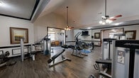 Located at 244 Kyser Blvd in Madison, Alabama, this single-floor fitness facility offers a comprehensive workout environment spread across three dedicated exercise rooms and two additional spaces. The facility blends two distinct architectural styles: a charming vaulted section with a natural wood-paneled ceiling, ceiling fan, and large windows that flood the space with natural light, housing treadmills, elliptical machines, and a stationary bike on tile flooring; and a more traditional interior zone featuring hardwood-style flooring, wainscoting with black trim accents, vaulted and tray ceilings, dual ceiling fans, and wall-mounted televisions. The strength training areas are thoroughly equipped with a wide array of selectorized machines, cable systems, free-weight benches, and a well-stocked dumbbell rack. Full-length mirrors line portions of the walls, enhancing the sense of openness and allowing users to monitor their form. Framed artwork and decorative touches add a polished, welcoming ambiance throughout. The interconnected layout of the rooms allows for a seamless flow between cardio, strength, and functional training zones, making this a versatile and well-appointed fitness destination.