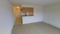 This single-floor unit offers a straightforward, functional layout centered around an open living space and an integrated kitchen. The living area is finished with wall-to-wall carpet in a neutral beige tone, creating a soft underfoot feel and contributing to the overall understated color palette throughout the space.

The kitchen is positioned at the rear of the main living area, separated by a half-wall with a built-in countertop that serves as a natural divider between the two spaces. Upper cabinetry with a warm wood-toned finish provides storage, while the kitchen itself is equipped with a gas range and a refrigerator. The kitchen floor transitions to tile, offering a practical, easy-to-clean surface in the cooking and entry zones.

A hallway leads from the kitchen area toward the entry door, which is illuminated by a ceiling-mounted light fixture, ensuring the transition space remains well-lit. The walls throughout are finished in a clean white, maintaining a bright and neutral atmosphere. Overall, the space presents a simple, no-frills design that prioritizes practicality and efficient use of its available footprint.