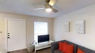 This single-floor space offers a practical and comfortable layout suited for everyday living. The living area serves as the heart of the home, featuring warm wood-style flooring that adds a sense of warmth and continuity throughout the space. A textured ceiling with a brushed nickel ceiling fan provides both air circulation and ambient lighting, keeping the room comfortable year-round. Natural light filters in through a window fitted with horizontal blinds, creating a bright and welcoming atmosphere. The living area is well-appointed, with ample space for seating and entertainment, and the neutral wall tones provide a versatile backdrop that complements a range of décor styles. A low-profile media console positioned beneath the window offers a clean and organized setup for a television and media equipment. The overall ambiance of the space is casual and inviting, with a simple, uncluttered aesthetic that makes it easy to personalize. The thoughtful use of space across the single floor ensures that every area feels purposeful and accessible, making this a functional and comfortable place to call home.