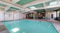 This indoor pool facility offers a generously sized swimming pool set within an architecturally distinctive enclosure. The ceiling is a standout feature, composed of painted wood planks and exposed beams in a muted green finish, accented by skylights that draw in natural light and brighten the water's surface. The pool itself is framed by a tiled deck with metal railings providing safe entry points into the clear, turquoise water. The surrounding walls combine smooth painted surfaces with textured wood paneling, contributing to a warm, enclosed atmosphere. Along the perimeter, a dedicated lounge and seating area features tables and chairs, offering a comfortable space for relaxation before or after a swim. Wall-mounted lighting fixtures supplement the natural illumination from the skylights, ensuring the space is well-lit throughout. An access corridor with doors is visible at the far end, providing connectivity to adjacent amenities or changing facilities. The overall design balances functional utility with a comfortable, inviting ambiance, making this indoor pool an excellent resource for year-round aquatic use.