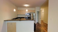This single-floor unit showcases a well-designed open-concept layout centered around a functional kitchen that flows seamlessly into the living areas. The kitchen is equipped with stainless steel appliances, including an electric range, built-in microwave, and a full-sized refrigerator. Light maple cabinetry lines the upper walls, offering generous storage, while dark granite countertops provide a striking contrast. A peninsula with a white base extends outward, offering additional counter space and serving as a casual breakfast bar. Pendant lighting hangs above both the peninsula and the main kitchen workspace, providing warm, focused illumination. The rich hardwood flooring extends throughout the open living area, adding warmth and visual continuity. A built-in open shelving nook is visible adjacent to the kitchen, offering a dedicated storage or display area. A hallway leads further into the unit, hinting at additional private spaces beyond. The overall design balances practicality with clean, modern aesthetics, creating an inviting and functional living environment.