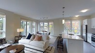 Located at 111 Morgan St in Stamford, Connecticut, this thoughtfully designed single-floor residence offers a seamless blend of modern aesthetics and functional living. The open-concept layout connects the living, dining, and kitchen areas into one cohesive space, making it ideal for both everyday living and entertaining. Large windows wrap around the main living area, flooding the interior with natural light and framing beautiful views of lush, mature trees that display vibrant autumn foliage. The living area features smooth, light-toned walls, dark hardwood flooring, and a clean white ceiling with track lighting, creating a bright and contemporary atmosphere. A plush area rug anchors the seating area, which opens naturally into a dining space positioned directly in front of expansive windows. The kitchen is defined by a sleek white peninsula with bar-height seating, pendant lighting, and what appears to be light stone countertops, offering a stylish and practical workspace. Throughout the space, the design palette is neutral and refined, with warm wood tones and crisp whites creating an inviting ambiance. The residence balances understated elegance with a welcoming warmth, enhanced by the greenery visible from nearly every angle.