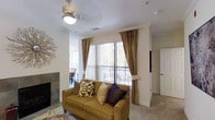 This single-floor residence offers a comfortable and thoughtfully designed living environment. The living area serves as the heart of the space, centered around a sofa and accented by a tiled fireplace that adds warmth and character. Large sliding glass doors flanked by elegant floor-length drapes allow generous natural light to fill the room, while vertical blinds offer privacy and light control. Views of lush greenery beyond the windows enhance the sense of connection to the outdoors. A ceiling fan overhead ensures year-round comfort. The neutral carpet and soft, warm color palette throughout create a cohesive and inviting ambiance. An open sightline connects the living area to a dining space, which also benefits from natural light streaming in through additional windows. A wall-mounted television and a conveniently placed interior door hint at the functional layout throughout the rest of the home. Crown molding details along the walls and ceiling add a refined architectural touch. Overall, this residence balances practical design with comfortable living, making it a welcoming and well-lit space suited for everyday life.