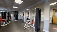 Located at 44 Brittany Farms Road in the Pebblebrook community of New Britain, Connecticut, this fitness center offers a comprehensive, single-floor workout facility designed to support a wide range of exercise routines. The gym features a spacious, open layout with a durable rubber flooring surface ideal for both cardio and strength training activities.

The space is outfitted with a solid selection of cardio equipment, including True-brand treadmills and elliptical machines, arranged in an orderly row to maximize usability and flow. A variety of strength training machines are distributed throughout the floor, including cable stations, a leg press, a shoulder press, and additional resistance machines with red-accented seats, accommodating full-body workout options.

Structurally, the room features white-painted support columns with wainscoting detail along the lower walls, painted in a two-tone gray scheme that lends a clean, well-maintained aesthetic. Floor-to-ceiling mirrors line one wall, visually expanding the space and allowing users to monitor their form during exercise. Recessed fluorescent panel lighting in a drop-tile ceiling provides consistent, even illumination throughout. Framed anatomical and motivational artwork decorates the walls, adding a purposeful, health-focused atmosphere. A water cooler station is also available for convenience.