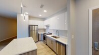 This contemporary single-floor unit offers a thoughtfully designed open-concept layout that seamlessly connects the living and kitchen areas. The kitchen is a standout feature, outfitted with high-gloss white upper cabinetry, warm wood-toned lower cabinets, and gleaming quartz countertops. A full suite of stainless steel appliances — including a refrigerator, range, and dishwasher — is complemented by a deep undermount sink with a professional-style pull-down faucet. Sleek pendant lighting and recessed ceiling lights add both style and functionality. A kitchen island provides additional prep and seating space, while light hardwood-style flooring extends throughout the main living areas, creating a bright and cohesive aesthetic. The living area is generously proportioned, featuring neutral gray walls, large sliding glass doors that invite natural light, and direct access to an outdoor area. The bathroom is designed with accessibility in mind, featuring a barrier-free walk-in shower with built-in grab bars, a fold-down bench, and clean large-format tile work alongside a wood-toned vanity with a white countertop. A spacious walk-in closet with wire shelving provides ample storage, completing this well-appointed and functional residence.