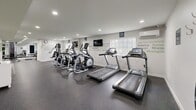Located in Glen Burnie, Maryland, this fitness center offers a thoughtfully designed exercise space that caters to a range of workout needs. The single-room layout is finished with durable rubber flooring in a speckled dark gray tone, providing both safety and functionality underfoot. Crisp white walls are adorned with bold motivational typography in green and gray, creating an energizing atmosphere throughout the space. Recessed lighting illuminates the room evenly, complementing the natural light that filters in through multiple windows. The cardio zone is well-stocked with treadmills, elliptical trainers, and a stationary bike, while a multi-station cable machine and a dedicated free weights rack serve strength training needs. An adjustable workout bench adds further versatility. A large mirror along one wall expands the sense of space and supports proper form during exercise. A wall-mounted flat-screen TV provides entertainment during workouts, and a water dispenser ensures convenient hydration. A wall-mounted air conditioning unit keeps the environment comfortable year-round. Hook-style coat storage and clearly posted safety and hygiene signage round out the practical amenities, making this a clean, organized, and motivating fitness facility.