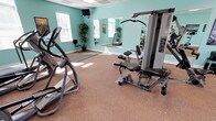 This fitness center offers a thoughtfully arranged workout space designed to accommodate a full range of exercise routines. The room is defined by its distinctive teal-painted walls and neutral-toned carpeted flooring, creating an energizing yet comfortable atmosphere. A drop-panel ceiling with recessed lighting panels and ceiling fans ensures bright, even illumination throughout, while multiple windows allow natural light to fill the space.

The facility is well-stocked with a diverse selection of equipment. Cardio options include treadmills, elliptical trainers, and an upright stationary bike, strategically positioned to take advantage of natural light from the windows. A multi-station cable weight machine serves as a centerpiece for strength training, complemented by a dedicated free-weight area featuring a tiered dumbbell rack with a full range of weights and a flat workout bench. Interlocking foam floor mats provide a designated stretching and floor exercise zone.

Large wall-mounted mirrors span one side of the room, visually expanding the space and allowing users to monitor their form during workouts. Motivational posters and decorative potted plants add character to the environment. A wall-mounted television provides entertainment during workouts, and a small utility shelf offers convenient storage for accessories.