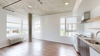 This 2-bedroom, 1-bathroom unit showcases a striking industrial aesthetic defined by exposed concrete ceilings and a prominent structural concrete column in the open living area. Rich hardwood flooring runs throughout the main living and kitchen spaces, complemented by an abundance of natural light streaming through multiple large windows that frame sweeping urban skyline and treetop views. The open-concept living area flows seamlessly into the kitchen, which features dark wood cabinetry, open shelving, a stainless steel range hood, and a full suite of white appliances including a refrigerator, dishwasher, and stove. Both bedrooms are carpeted for comfort and offer generous closet space behind sliding wood-panel doors, with each room benefiting from its own window with city views. The exposed concrete ceiling extends into the bedrooms, maintaining a cohesive industrial character throughout. A hallway connects the private areas of the unit, leading to the full bathroom, which includes a tub/shower combination, toilet, and a dark wood storage cabinet for added functionality. The clean white walls throughout provide a bright, neutral canvas that balances the raw industrial elements, creating a modern and practical urban living space.