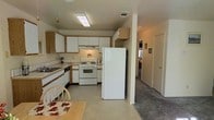 This single-story residence offers a practical and well-organized layout designed for comfortable everyday living. The kitchen serves as a central feature of the home, equipped with white cabinetry accented by wood-tone trim, a gas range, a full-size refrigerator, and a built-in dishwasher. A tile countertop with an integrated sink provides ample workspace, while overhead fluorescent lighting ensures the area is well-lit. Cabinet storage is plentiful, with both upper and lower units lining the walls to maximize functionality. A dining area sits adjacent to the kitchen, accommodating a wooden table and chairs, making it convenient for casual meals. The kitchen flows naturally into a hallway that leads to the rest of the home, where carpeted flooring creates a warmer, softer feel underfoot. The hallway connects to additional rooms, visible in the background, suggesting a straightforward and accessible floor plan throughout. Neutral wall tones and consistent finishes across the space contribute to a cohesive and understated aesthetic. Overall, the home presents a clean, functional environment well-suited for those seeking a straightforward and practical living arrangement on a single level.