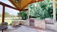This single-level outdoor pavilion is designed for comfortable al fresco living and entertaining. The structure is anchored by a steeply pitched, vaulted ceiling finished in warm wood tongue-and-groove planking, supported by sturdy timber beams and columns that lend a rustic yet refined character to the space. A ceiling fan is mounted at the apex, ensuring comfortable airflow throughout the warmer months.

The centerpiece of the pavilion is a generously sized built-in outdoor kitchen, constructed with natural stone masonry in a warm sandy hue. The kitchen counter wraps in an L-shape, providing ample prep and serving space, and is equipped with a stainless steel built-in grill with storage cabinetry below — ideal for outdoor cooking and entertaining.

The concrete flooring is durable and easy to maintain, suited for heavy outdoor use. Open on multiple sides, the pavilion offers unobstructed views of a lush, mature tree line that creates a private, park-like backdrop. A wrought iron fence is visible in the background, subtly defining the property boundary without interrupting the natural scenery. The overall ambiance is relaxed and inviting, blending functional outdoor cooking amenities with the natural beauty of the surrounding landscape.