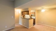 This single-floor residence offers a practical and comfortable layout with notable features throughout. The living area is a welcoming space anchored by a dark tile-surround fireplace, serving as a natural focal point. Soft carpeting, neutral-toned walls, and a large window fitted with blinds allow natural light to fill the room, contributing to a bright and open atmosphere. The vaulted ceiling adds a sense of vertical spaciousness to the area.

The kitchen is thoughtfully designed with an open-concept approach, connected to the living and dining areas via a raised breakfast bar with a dark countertop — ideal for casual dining or conversation. Warm wood-toned cabinetry lines the walls, providing ample storage, while white appliances including a refrigerator and range are neatly integrated into the layout. An overhead range hood and additional open shelving add both function and character to the space.

A ceiling fan is present in the adjoining area, enhancing comfort and air circulation. The consistent use of neutral wall colors and carpet throughout creates a cohesive, understated aesthetic. Overall, this space combines functional design with cozy residential touches, making it a practical and inviting single-floor home.