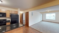 This single-floor residence offers a well-organized, open-concept layout that seamlessly connects the kitchen and living areas. The kitchen is equipped with light maple cabinetry, dark granite countertops, and a black refrigerator, providing both functionality and a clean aesthetic contrast. Hardwood-style flooring extends throughout the main living space, lending warmth and continuity to the open floor plan.

The living area transitions smoothly from the kitchen, featuring plush carpeting that adds a layer of comfort underfoot. Natural light filters in through a door with a glass panel at the far end of the living space, creating a bright and welcoming atmosphere. White walls throughout the space keep the interior feeling fresh and neutral.

Several classic six-panel interior doors in a rich wood finish open to additional rooms, suggesting a thoughtful room count for the floor plan. A hallway branching off the main area provides access to these private spaces. HVAC vents are integrated into the walls, ensuring efficient climate control throughout. The textured ceiling and warm accent walls add subtle visual interest, making this a comfortable and practical single-level living space.