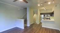This two-story residence showcases an open-concept main level where the living area flows seamlessly into the kitchen. Rich, dark hardwood-style flooring runs throughout the main floor, providing a warm contrast to the light-toned walls and white trim. The living area is equipped with a ceiling fan, while track lighting overhead in the kitchen area ensures well-lit functionality.

The kitchen features dark cabinetry, stainless steel appliances including a full-size refrigerator, and a light-colored countertop peninsula that serves as both a workspace and a casual dining or gathering surface. A tile backsplash adds a decorative touch to the kitchen backdrop.

Two stairways are visible from the main level — one ascending and one descending — indicating that the living space spans multiple floors, offering separation between living and private areas. Crown molding details along the ceiling add a refined architectural touch throughout the main level. The overall ambiance is modern and practical, with clean lines, neutral tones, and thoughtful design elements that create a comfortable and cohesive living environment.
