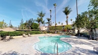 This outdoor recreational area offers a well-appointed resort-style escape designed for relaxation and leisure. The centerpiece is a generously sized, freeform swimming pool with a smooth stone surround and stainless steel entry rails, its clear turquoise water reflecting the abundant California sunshine. Adjacent to the pool sits a circular spa, perfectly positioned for unwinding, set within an elegant stone patio finished in large-format pavers that lend a clean, upscale aesthetic to the space.

Towering queen and royal palm trees frame the area dramatically, creating a tropical ambiance and providing natural shade throughout the day. Manicured hedgerows line the perimeter, offering a sense of privacy and enclosure while still allowing views of the surrounding landscape and neighboring buildings. Lounge chairs are thoughtfully placed along the deck, inviting residents to soak in the sun or relax poolside.

The overall atmosphere is bright, open, and inviting, with lush greenery softening the hardscape elements. The combination of tropical landscaping, clean stonework, and well-maintained water features creates a cohesive outdoor environment that evokes a serene, resort-like setting ideal for both quiet relaxation and casual social gatherings.
