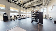 This two-floor fitness facility offers a comprehensive and well-equipped training environment across a generously sized footprint. The main floor welcomes visitors with warm hardwood flooring at the entry, glass-partitioned walls, and dramatic motivational wall graphics that set an energetic tone. A wide indoor sprint track with marked lanes runs prominently through the open training floor, serving as the centerpiece of the functional training zone. Surrounding the track, racks stocked with kettlebells, medicine balls, resistance bands, and plyo boxes provide everything needed for high-intensity workouts. A staircase connects the two levels, with the upper floor housing a dedicated cardio zone lined with treadmills, stationary bikes, and ellipticals positioned alongside panoramic windows that flood the space with natural light and offer expansive outdoor views. Exposed ductwork and industrial-style ceilings with track and pendant lighting give the upper level a modern, loft-like aesthetic. A separate strength training area features a full array of cable machines, benches, and free weights beneath soaring ceilings. A private workout room finished in warm wood paneling features a smart fitness mirror and natural light, ideal for personal training or focused sessions. Throughout, rubber flooring, open sightlines, and abundant natural light create an inviting, performance-oriented environment.