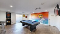 Located at 7000 Phoenix Ave NE in Albuquerque, New Mexico, this single-room game room is a lively and well-appointed entertainment space designed for leisure and social activity. The room is anchored by a full-size pool table with a blue felt surface, positioned centrally on stylish herringbone-patterned hardwood-look flooring that adds a sophisticated visual texture to the space.

One of the most striking features is a bold, floor-to-ceiling mural depicting a colorful hot air balloon soaring against a vivid sunset sky with billowing clouds — a nod to Albuquerque's renowned ballooning culture. This eye-catching wall art lends the room an energetic and playful atmosphere.

Additional recreational amenities include a full arcade dart board cabinet and a wall-mounted flat-screen television, making the space suitable for a variety of entertainment activities. A row of bar-height stools lines one wall alongside a large, colorful abstract artwork, providing casual seating for spectators or guests. Decorative signage adds personality and charm throughout.

The room is finished with clean white walls, recessed lighting, and ample HVAC venting to ensure comfort. A large window allows natural light to filter in, while an adjoining door provides access to adjacent areas. Overall, the space delivers a fun, welcoming environment tailored for community recreation.