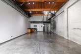 Located in Los Angeles, this two-floor industrial loft at 201 N Westmoreland Ave blends raw character with modern functionality across a generous layout. The main level makes an immediate impression with polished concrete floors, exposed concrete block walls, and striking wood-beam ceilings fitted with track lighting. A dramatic black spiral staircase serves as a sculptural centerpiece, connecting the ground floor to the upper loft. The open-plan kitchen and living area on the main floor features warm wood cabinetry, dark granite countertops, stainless steel appliances, and exposed structural columns that reinforce the space's industrial aesthetic. Glass block windows allow natural light to filter through while maintaining privacy. The upper loft level offers a refined contrast, with rich hardwood flooring, crisp white walls, a ceiling fan, and expansive glass garage-style doors that flood the space with natural light and provide outdoor views. Exposed ductwork and pipe detailing carry the industrial theme throughout. The loft level also features a sleek stainless cable railing along the open stairwell. Two bathrooms serve the space. The overall ambiance is bold and characterful — a versatile urban environment that balances raw, warehouse-inspired design with thoughtful modern finishes.