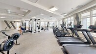 Located at 320 Fisher St in Charlottesville, Virginia, this well-appointed fitness center at Altoview Apartments is a single-floor facility designed to support a comprehensive workout experience. The space is generously sized, offering dedicated zones for cardio training, strength equipment, and functional fitness. A long row of treadmills, ellipticals, and stationary bikes lines the perimeter, each positioned to take full advantage of the large windows that flood the room with natural light and offer direct views of the outdoor pool area. The strength and functional training section is equally well-equipped, featuring a multi-station cable rack, weight benches, barbells, medicine balls, resistance bands, a heavy bag, and rowing machines. A dedicated room visible through an interior doorway provides additional space for stretching or focused exercise, complete with a large display screen. Throughout the facility, bold geometric diamond and chevron wall graphics in dark tones add a modern, energetic aesthetic against the neutral palette of light gray carpet and white ceilings. Ceiling fans and recessed panel lighting ensure a comfortable environment. The overall design is clean, bright, and motivating, reflecting a high-quality amenity space suited for a wide range of fitness routines.