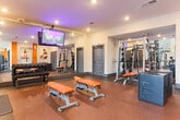 Located at 151 Integra Vistas Dr in Chattanooga, TN, this fitness center offers a thoughtfully designed workout environment across a single floor. The space features a contemporary aesthetic defined by warm orange accent walls, neutral tones, and speckled rubber flooring throughout, providing both style and functionality.

The main training area is generously proportioned, accommodating a wide variety of equipment including selectorized strength machines, free weight benches with orange upholstery, dumbbell racks, a suspension training rig, and cable machines. Large wall-mounted mirrors amplify the sense of space and allow users to monitor their form during workouts. Recessed lighting and ceiling-mounted fixtures ensure the space is well-lit throughout.

A ceiling-mounted flat-screen television is positioned prominently for visibility across the workout floor. Fitness-themed artwork decorates the walls, adding motivational character to the environment. A staircase with glass railings and wood treads connects to an upper level, accompanied by a decorative pendant light fixture that adds an upscale touch. Clerestory windows near the staircase bring in natural light, enhancing the open, airy feel of the space. Additional rooms accessible via dark-paneled doors offer supplementary storage or utility functions. The overall ambiance is clean, modern, and purpose-built for comprehensive fitness training.