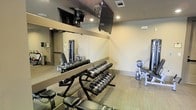 This fitness room is designed to deliver a fully functional workout environment within a single-level layout. The space is outfitted with a tiered dumbbell rack holding a wide range of free weights, providing options for both light and heavy resistance training. A cable weight machine is positioned along the mirrored wall, expanding the variety of exercises available to users. Additional strength equipment, including a weight bench and plate-loaded machine, can be seen toward the far end of the room, making efficient use of the available floor space.

The mirrored wall along one side visually expands the room while allowing users to monitor their form during workouts. Wall-mounted flat-screen televisions are installed near the ceiling, offering entertainment during exercise sessions. Recessed lighting keeps the space well-lit throughout. The neutral-toned walls and carpeted flooring contribute to a clean, low-distraction environment suited for focused training. Ventilation is provided via ceiling vents, supporting comfort during intensive workouts. Overall, the fitness room is thoughtfully arranged to balance equipment variety with ease of movement.