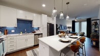 This well-designed single-floor residence combines modern aesthetics with functional living. The open-concept kitchen is a standout, featuring crisp white shaker cabinetry, a bold blue herringbone tile backsplash, white quartz countertops, and a full suite of stainless steel appliances including a range, dishwasher, microwave, and refrigerator. A central kitchen island with pendant lighting overhead provides additional seating and workspace, flowing naturally into a dedicated dining area. The living room offers rich hardwood-style flooring, large windows with floor-to-ceiling drapes that invite generous natural light, a ceiling fan, and direct access to what appears to be an exterior door, lending an open, airy feel to the space. The bedroom is a calm retreat, highlighted by a deep teal accent wall, plush carpeting, and wide windows that fill the room with soft, natural light. The primary bathroom is well-appointed with a dual-sink vanity featuring grey cabinetry and white countertops, hardwood-style flooring, open shelving for storage, and a spacious walk-in closet with built-in organization. Throughout the residence, a cohesive contemporary design palette of whites, greys, and bold accent tones creates a polished and inviting atmosphere.
