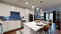 This well-designed single-floor residence combines modern aesthetics with functional living. The open-concept kitchen is a standout, featuring crisp white shaker cabinetry, a bold blue herringbone tile backsplash, white quartz countertops, and a full suite of stainless steel appliances including a range, dishwasher, microwave, and refrigerator. A central kitchen island with pendant lighting overhead provides additional seating and workspace, flowing naturally into a dedicated dining area. The living room offers rich hardwood-style flooring, large windows with floor-to-ceiling drapes that invite generous natural light, a ceiling fan, and direct access to what appears to be an exterior door, lending an open, airy feel to the space. The bedroom is a calm retreat, highlighted by a deep teal accent wall, plush carpeting, and wide windows that fill the room with soft, natural light. The primary bathroom is well-appointed with a dual-sink vanity featuring grey cabinetry and white countertops, hardwood-style flooring, open shelving for storage, and a spacious walk-in closet with built-in organization. Throughout the residence, a cohesive contemporary design palette of whites, greys, and bold accent tones creates a polished and inviting atmosphere.