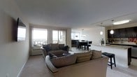 This single-floor space offers an open-concept layout that seamlessly connects the living and dining areas, creating a fluid and functional environment. The living room is generously proportioned, anchored by a large, plush sectional sofa in warm taupe tones and complemented by coordinating accent pillows. Soft, neutral-toned carpeting runs underfoot, contributing to the comfortable and inviting ambiance throughout the space. Large windows and a sliding glass door flood the living area with abundant natural light, while offering expansive views of the surrounding cityscape and beyond — a standout feature of this space. The dining area transitions naturally from the lounge, centered around a dark-toned table and chairs beneath elegant pendant lighting, adding a refined touch to the open floor plan. A partial wall with a pass-through glimpse into an adjoining room hints at a kitchen or additional living space nearby. The overall design aesthetic is contemporary and understated, with a cohesive neutral color scheme, clean lines, and a blend of comfort and sophistication that makes the space both practical and visually appealing.