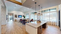 Located at 19815 Kenswick Drive in Humble, Texas, this single-story community space showcases a thoughtfully designed open-plan layout that seamlessly connects multiple functional zones. Light hardwood-style flooring runs throughout, providing a warm, cohesive foundation that complements the crisp white walls and vaulted ceilings. Clerestory windows positioned along the upper walls invite abundant natural light while maintaining privacy, lending the interior a bright and airy atmosphere.

The welcoming lounge area features vaulted ceilings accented by exposed wood beam details and is furnished with plush wingback chairs, sofas, and round coffee tables arranged to encourage conversation. Built-in shelving niches and a wall-mounted flat-screen television add functional character to the seating zone. Large windows at the far end offer views of the outdoors, enhancing the sense of openness.

Adjacent to the lounge, a well-appointed kitchenette serves as a central gathering point. A generously sized island with a quartz countertop and bar seating anchors the space, while statement pendant lights hang above in a lantern style. A textured tile accent wall frames the countertop workspace, which includes a sink, under-counter refrigeration, and ample storage with dark cabinetry. The overall ambiance is modern, polished, and welcoming.