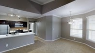 This single-floor residence offers a thoughtfully designed open-concept layout that seamlessly connects the living, dining, and kitchen areas into one cohesive space. The living and dining areas are finished with warm, neutral carpeting that adds comfort underfoot, while the walls are painted in a sophisticated gray tone complemented by crisp white crown molding that runs throughout, adding a refined architectural detail to each room.

The kitchen is a standout feature, showcasing sleek dark cabinetry that contrasts beautifully against light countertops and stainless steel appliances, including a range and microwave. A white kitchen island or peninsula extends into the open area, providing additional prep space and defining the transition between the kitchen and living spaces.

Natural light filters into the dining area through a window dressed with blinds, while a contemporary pendant light fixture hangs above, serving as a focal point and providing warm illumination. The tray ceiling detail in the main living area adds visual depth and a sense of elevated design. Hallways visible from the main space suggest additional rooms beyond, contributing to the overall functional flow of the floor plan. The neutral, modern palette and open layout create a bright, welcoming ambiance throughout.