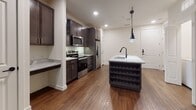 This thoughtfully designed single-floor residence offers a cohesive blend of modern finishes and functional living spaces. The kitchen serves as the heart of the home, showcasing rich dark cabinetry that contrasts beautifully with light granite countertops featuring subtle veining. A central island with a built-in wine rack and undermount sink is illuminated by sleek pendant lighting, creating an inviting focal point. Stainless steel appliances, including a French door refrigerator, electric range, and microwave, complete this well-appointed culinary space. Warm hardwood-style flooring flows throughout the main living areas, adding depth and continuity to the open layout. The bathroom is finished with refined details, including a dark-framed vanity mirror, a vessel-style sink atop a granite countertop, dark wood cabinetry with brushed hardware, and matte black fixtures that lend a contemporary edge. Dark tile flooring anchors the space with a clean, modern aesthetic. The bedroom features plush neutral carpeting underfoot and is bathed in natural light from a pair of windows dressed with horizontal blinds. A ceiling fan adds comfort and practicality. The overall ambiance throughout the space is clean, modern, and move-in ready, with a neutral palette that offers versatility for a variety of interior styles.