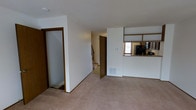 This three-floor space offers a functional and straightforward layout with clean, neutral interiors throughout. The main level is anchored by a spacious carpeted area that serves as the central living space, finished with white walls and warm wood-toned door frames and trim that add a touch of contrast to the otherwise light palette.

A defining feature of the main level is the semi-open kitchen, separated from the living area by a half-wall with a pass-through counter. The kitchen is equipped with dark appliances, including a refrigerator, and cabinetry with wood-toned finishes. A small window above the counter allows natural light to filter into the kitchen area.

The layout includes multiple doorways leading to additional rooms and access points. A staircase visible from the main level connects to the upper and lower floors, facilitating easy movement throughout all three levels. A floor vent is integrated into the main living area, indicating a central heating and cooling system.

Overall, the space presents a clean, neutral, and move-in ready environment with a practical multi-level design suited for everyday living.