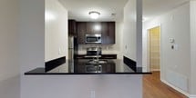 This single-floor residence combines modern design with functional living spaces. The kitchen is a standout feature, showcasing rich dark espresso cabinetry, striking dark granite countertops, a neutral tile backsplash, and a full suite of stainless steel appliances including a refrigerator, over-the-range microwave, and electric range. A peninsula with a waterfall granite countertop opens directly to the living area, creating a seamless flow ideal for both everyday living and entertaining. The living room is generously sized, finished with warm hardwood-style flooring and neutral-toned walls, and receives natural light through a large window. The bedrooms are carpeted in a soft, neutral tone, offering comfort underfoot. One bedroom features a built-in closet organizer with shelving and hanging space, providing ample storage. The bathrooms are thoughtfully finished; one includes a dark vanity with a granite countertop and a tiled walk-in shower, while the second offers a lighter vanity with a mirrored cabinet and tile flooring. Throughout the space, consistent use of dark cabinetry hardware and neutral wall tones creates a cohesive, contemporary aesthetic.