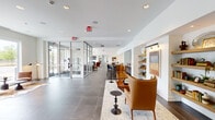 Located on Ellinwood Street in Des Plaines, Illinois, this thoughtfully designed single-floor amenity space offers a seamless blend of modern aesthetics and functional comfort. The layout extends along a generous corridor anchored by large-format gray tile flooring throughout, creating a cohesive and polished foundation for the various zones within.

The entrance features floor-to-ceiling glass partition walls with aluminum framing, allowing natural light to flow freely while defining distinct functional areas. A welcoming lounge area near the entry is furnished with rich cognac leather chairs, a sculptural chandelier, and oversized windows that frame views of the surrounding streetscape, lending an open, airy quality to the space.

Moving deeper into the interior, a dining and social zone features leather banquette seating with marble-top tables and nailhead-trim accent chairs, illuminated by warm wall sconces set against textured panel backdrops. A communal high-top table with blue bar seating provides an additional gathering option nearby.

A wet bar kitchenette is outfitted with warm wood cabinetry, a light quartz countertop, a tile backsplash, a stainless steel sink, and a beverage refrigerator — ideal for hosting and entertaining. Built-in shelving with curated décor and artwork further enhances the refined, residential-inspired atmosphere throughout this well-appointed community space.