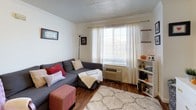 This single-floor unit offers a well-organized layout that makes efficient use of its space across key living areas. The living room serves as the heart of the unit, featuring hardwood flooring, crown molding, and a large window dressed with sheer white curtains that allow natural light to fill the room. A wall-mounted HVAC unit provides climate control, and the space comfortably accommodates a sectional sofa and open shelving.

The kitchen is designed for practicality, featuring light wood cabinetry with black hardware, white laminate countertops, and tile flooring. It is equipped with an electric range, dishwasher, and refrigerator, offering ample cabinet and counter space for daily meal preparation.

The unit includes two bedrooms, both carpeted and naturally lit through windows with warm-toned curtains and blinds. Each room provides a comfortable and personal retreat, with ceiling fixtures and space for storage furniture. The rooms share a neutral color palette that lends a calm, understated atmosphere.

The bathroom is straightforward and functional, featuring tile flooring, a single vanity with storage cabinetry, a toilet, and a shower-tub combination enclosed by a curtain. The layout is clean and easy to navigate. Overall, the unit presents a practical and livable space suited for individuals or small households.