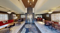 Located at 1500 Plantation Oaks Drive in Trophy Club, Texas, this well-appointed clubhouse offers a refined and welcoming environment designed for community use. The space is organized across a single floor, encompassing a living room and a dining room that together form a cohesive, functional gathering area.

The entry corridor immediately impresses with its dramatic vaulted ceiling, accented by a warm brown tray detail and an elegant chandelier that draws the eye upward. Dark-toned French doors with glass panels serve as a distinguished focal point, flanked by interior glass partition walls that visually connect the spaces while maintaining definition between zones.

The living room features rich, dark wall tones complemented by woven window shades that filter natural light. Seating includes a bold red sofa, upholstered armchairs, a rustic wooden coffee table, and round ottomans arranged around a patterned area rug — creating an inviting, lounge-like atmosphere. A ceiling fan and recessed lighting provide comfortable illumination throughout.

The dining area is equally well-equipped, offering a sturdy wooden dining table with mixed seating and a dedicated workstation with dual monitors, making it suitable for both social gatherings and productive work sessions. Woven shades, decorative wall art, and a ceiling fan contribute to the room's warm, grounded aesthetic. Tile flooring flows consistently throughout the entire space, tying the design together with a clean, polished finish.
