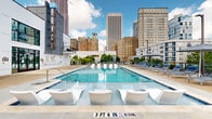 Located at 131 Ponce de Leon Ave NE in Atlanta's vibrant Midtown neighborhood, this outdoor amenity deck offers a compelling blend of urban energy and resort-style relaxation. The centerpiece is a generously sized swimming pool with crystal-clear turquoise water, flanked on all sides by a broad concrete sundeck. Rows of cushioned lounge chairs and market umbrellas provide ample space for sunbathing, while in-pool sun ledges with contemporary white chaise loungers invite residents to relax at the water's edge. The deck also features a covered outdoor bar and lounge area equipped with a marble-topped counter, bar stools, mounted flat-screen televisions, and ceiling fans, making it an ideal setting for casual gatherings. High-top tables with bar seating are positioned along the perimeter, taking full advantage of the panoramic views of Atlanta's skyline, including several prominent high-rise towers that frame the horizon. The surrounding architecture reflects a modern residential aesthetic, with glass curtain walls, clean panel facades, and private balconies visible on the adjacent building. Lush greenery along the fencing softens the urban landscape, while open skies above ensure abundant natural light throughout the day.