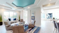 This thoughtfully designed single-floor amenity space blends contemporary elegance with functional comfort across several distinct areas. The central lounge is an immediate showpiece, featuring a dramatic coffered ceiling with a bold teal inset, recessed lighting, and large windows that flood the room with natural light and views of surrounding greenery. Taupe accent chairs with chrome frames, a round glass coffee table, and a bold blue-toned area rug anchor the space with refined coastal style. Adjacent sitting areas continue the sophisticated aesthetic, with gallery walls of framed abstract artwork in gold frames, plush neutral sofas accented with deep blue cushions, and light tile flooring throughout. The open kitchen is sleek and well-appointed, featuring white cabinetry, a large waterfall-edge island with seating for several, stainless steel appliances, double wall ovens, and a striking blue mosaic tile backsplash that frames a mounted flat-screen TV. Pendant lighting overhead completes the polished look. A dedicated game room offers a round poker table surrounded by elegant chairs, large windows with exterior views, and bold abstract canvas art on the walls. Throughout the entire space, a cohesive palette of crisp white, warm taupe, and coastal blue creates a bright, welcoming, and upscale atmosphere.